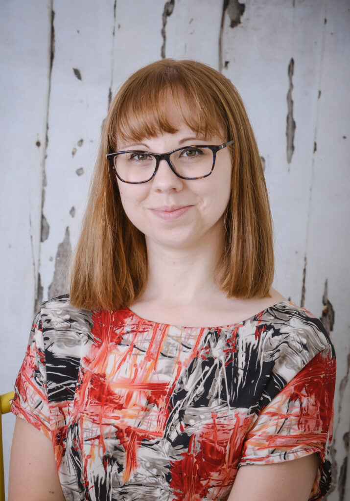A headshot of a woman in her 30s with light brown hair and square glasses. She is wearing a red, blank adn grey abstract print top and is in front of a rustic wood background